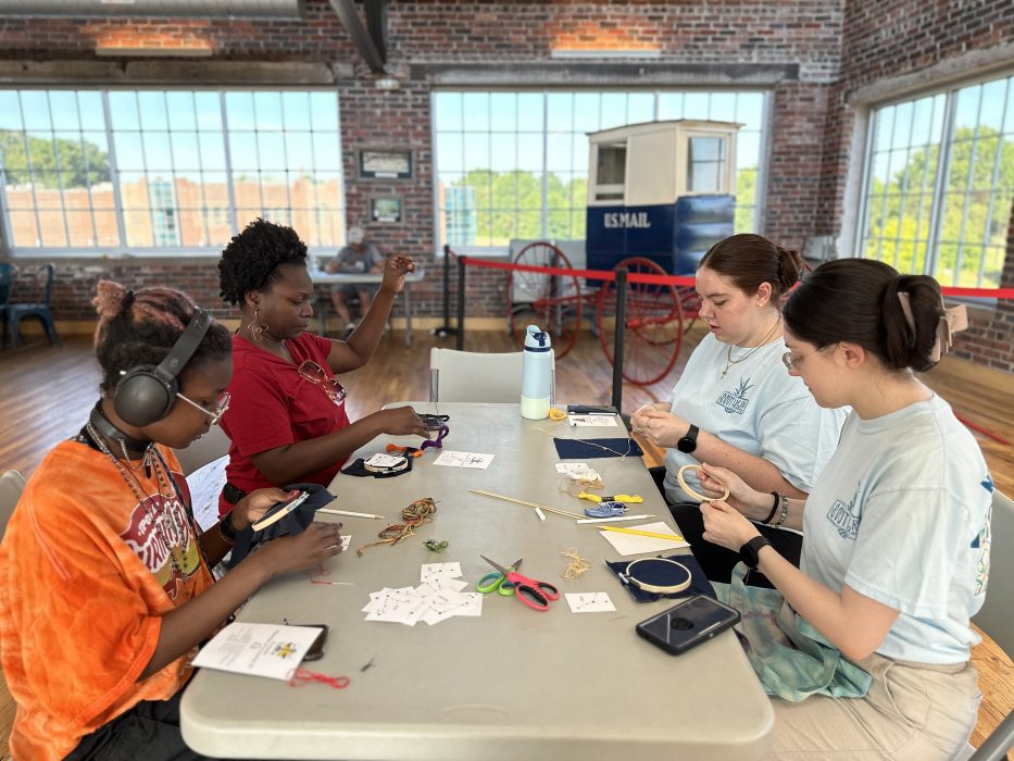 Four women sit around a table learning how to embroider a pattern.