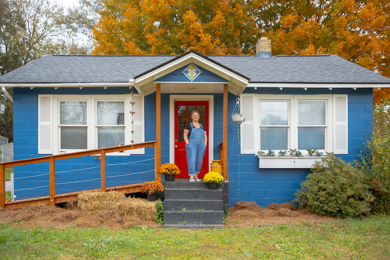 Stacey P Smith standing on the porch at The Blue House Studio, a brick home painted black.